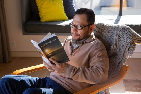 Mature asian man reading paperback endless sunsets in sunlit cozy armchair