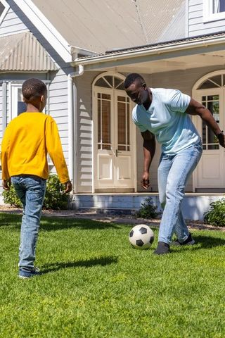 African american father coaching son while playing soccer on sunlit front lawn