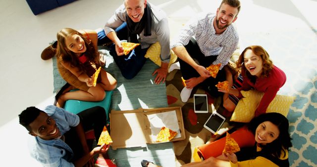 A diverse group of friends is sitting in a circle on the floor, smiling and enjoying slices of pizza from an open box on a cozy rug. They are chatting and laughing, surrounded by pillows and personal items such as tablets. This image can be used in contexts related to social gatherings, friendship, youth activities, parties, and casual dining.