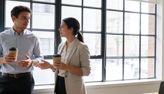 Two Colleagues Chatting over Takeaway Coffee by Large Grid Window in Bright Office