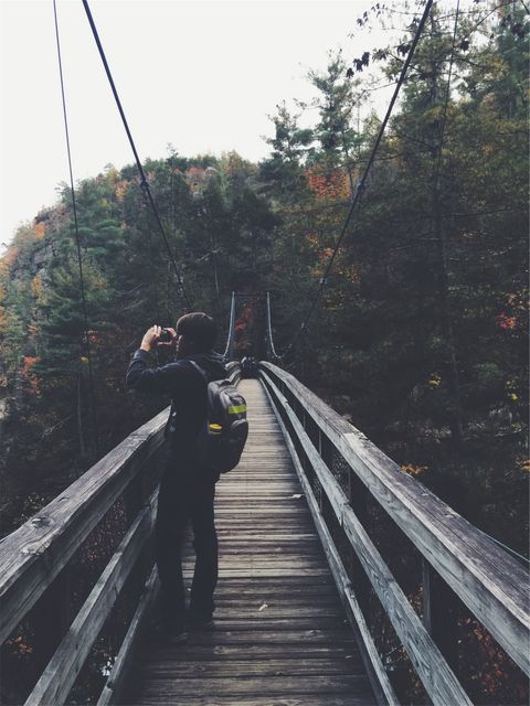 A person wearing a backpack photographing scenery on a secluded wooden suspension bridge in a dense forest. The bridge stretches over a forested ravine with trees showcasing fall colors in the background. Ideal for blogs or articles about outdoor adventures, travel destinations, and nature photography. Can also be used for promoting hiking gear, travel guides, and lifestyle content related to exploring remote areas.