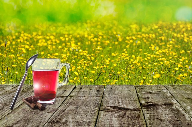 Red tea served in glass cup with spoon and cinnamon sticks on rustic table, overlooking a vibrant wildflower meadow under sunny sky. Ideal for illustrating outdoor relaxation, nature retreats, health and wellness concepts, springtime activities, and tea advertisements.