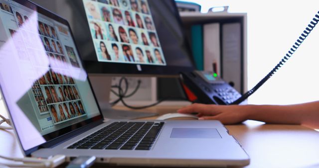 Person browsing a photo database on both a laptop and desktop monitor in an office, showing engagement in digital work. Great for themes like digital work, technology usage, office productivity, and online searches.