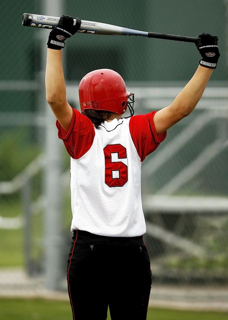 Player in red and white uniform raising bat above head, indicating celebration or victory in an intense softball moment. Great for use in articles about sports achievements, advertisements for sports gear, motivational posters, and promotional materials for softball events or training programs.