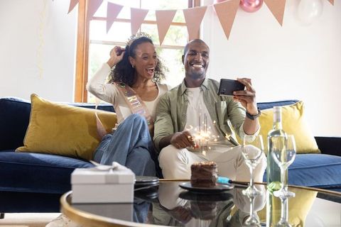 Diverse couple celebrating birthday at home taking selfie with cake, sparkler, wine