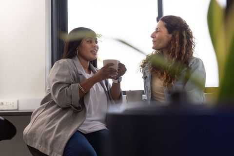 Diverse female coworkers sharing coffee and chatting in bright modern office lounge
