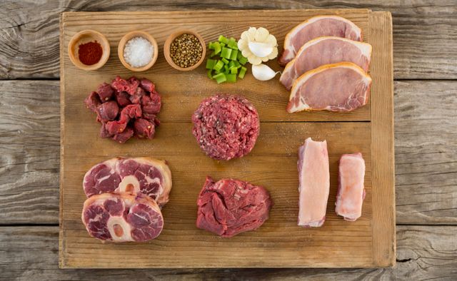 Assorted raw meats including beef, pork, and various cuts arranged on a wooden tray with spices such as garlic, salt, pepper, and paprika. Ideal for use in culinary blogs, cooking websites, butcher shop promotions, and food preparation guides.