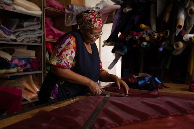 Side view of a biracial woman standing at a table working with purple fabric at a hat factory. In the background bales of material can be seen in storage.