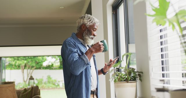 Senior man standing by window with natural light, drinking coffee and checking smartphone. Suitable for themes like daily routine, technology in mature lifestyles, relaxation at home, and indoor modern living.