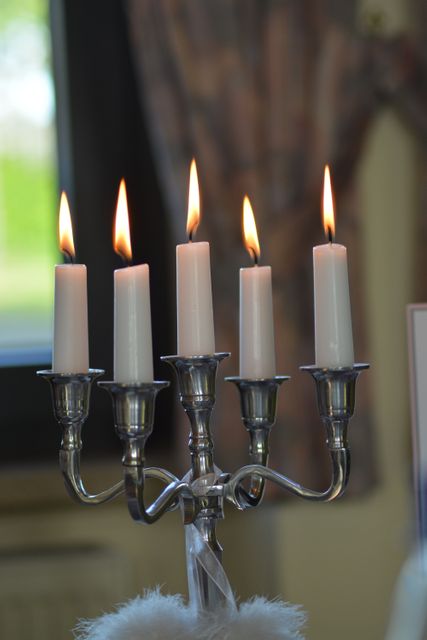 Five white candles burning on classic silver candelabra, creating warm ambiance in cozy room. Flames casting soft light, perfect for romantic dinners, festive gatherings, and elegant decors. Adds touch of vintage charm to any space.