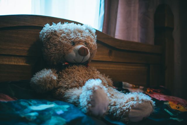 Teddy bear sitting on wooden bed in a softly lit room. This evokes feelings of comfort and nostalgia, ideal for use in content about childhood memories, children's room decoration ideas, or themes of coziness and warmth.