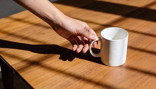 Hand grasping white ceramic mug on sunlit wood table casting window grid shadows