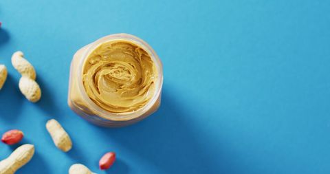Creamy peanut butter jar surrounded by peanuts on blue background