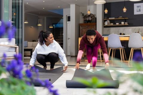 Rolling out yoga mats together: friends preparing sunrise home workout by open glass doors