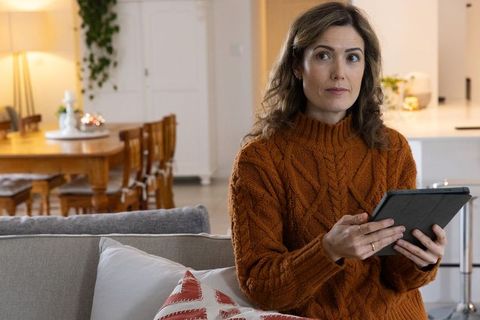 Woman wearing rust cable knit sweater holding tablet sitting on cozy living room sofa