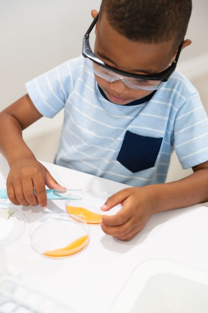 Young African American boy wearing safety goggles conducting a chemistry experiment in a laboratory. He is handling a test tube and petri dish, demonstrating hands-on learning in STEM education. Ideal for educational materials, science programs, school projects, and promoting STEM activities for children.