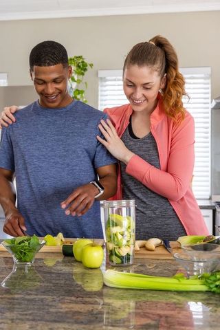 Couple preparing healthy green smoothie on kitchen island with apples, celery, spinach