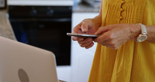 Close-up of elderly person in yellow dress using smartphone and laptop in kitchen. Image can be used to depict seniors interacting with technology, promoting digital literacy, and modern lifestyle for older adults.