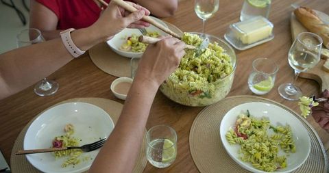 African american friends serving pasta salad and sharing meal around wooden dining table