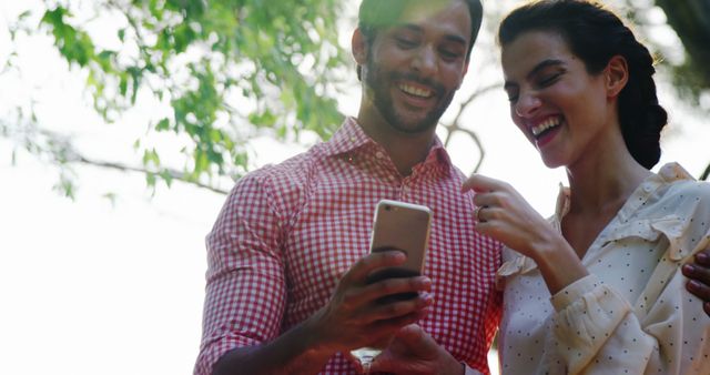 Young couple happily engaging with a smartphone, surrounded by greenery. This image is perfect for depicting modern relationships, happiness, and outdoor activities. It can be used in promotions for technology, lifestyle blogs, social media campaigns, or advertising outdoor products.
