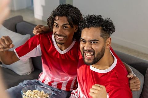 Cheering friends wearing red soccer jerseys on couch with popcorn and high energy celebration