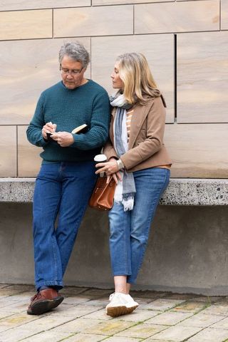 Senior couple resting against city bench, man jotting in notebook, woman holding coffee cup