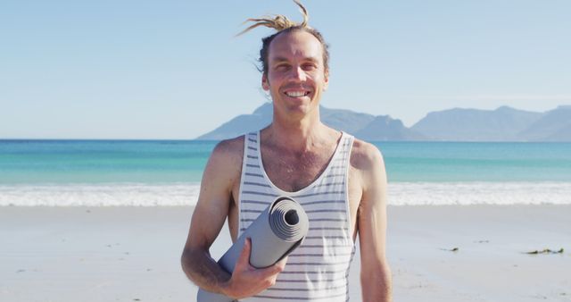 Man holding yoga mat at beachside smiling at camera. Ideal for themes around fitness, wellbeing, relaxation, and vacation. Perfect for blogs, travel websites, wellness programs, and outdoor adventure promotions.