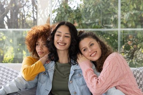 Diverse friends cuddling and smiling on sunlit sofa in glass sunroom enjoying bonding