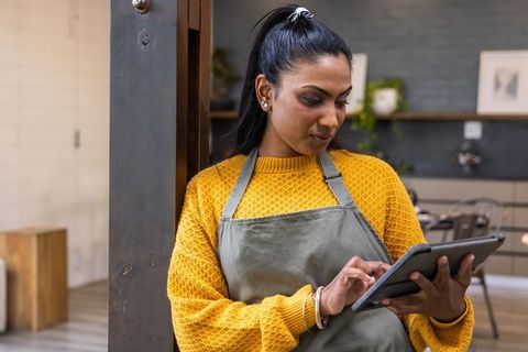 Indian cafe barista using tablet while leaning on wooden post in modern coffeehouse