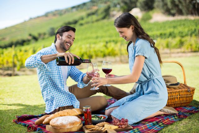 Couple enjoying a picnic in a vineyard, with man pouring red wine into glasses. Ideal for use in advertisements for wine brands, travel destinations, outdoor activities, and lifestyle blogs. Perfect for promoting romantic getaways, vineyard tours, and picnic products.
