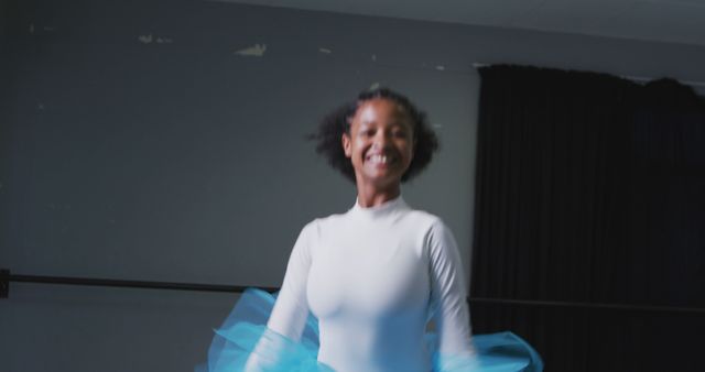 A young woman is smiling and practicing ballet in a studio. She is wearing a blue tutu and appears to be in mid-movement, capturing the joy and elegance of dance. This visual can be used to highlight themes of joy, dedication, and the arts. It is suitable for websites or magazines focused on ballet, dance schools, or performing arts promotion.