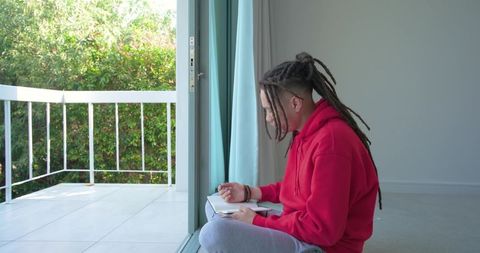 Mid-adult man with dreadlocks writing in notebook by sliding door sunny minimalist balcony