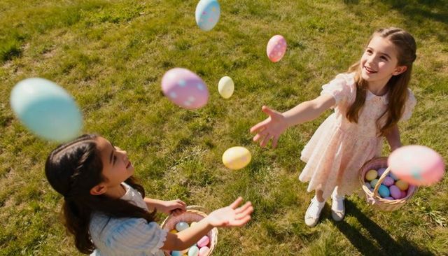 Two girls tossing pastel easter eggs during spring egg hunt on sunny lawn