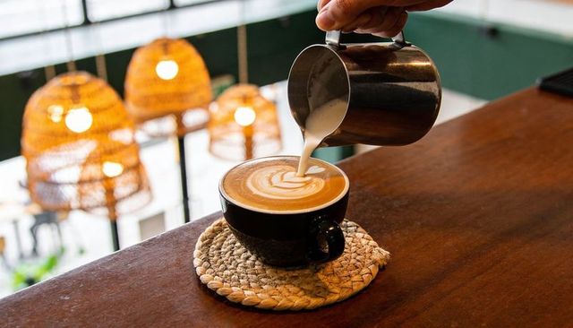 Barista pouring latte art in black cup on woven coaster with warm cafe lighting