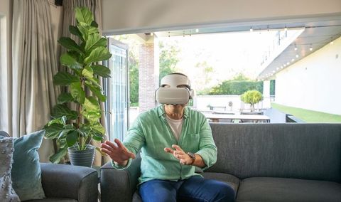 Mature woman wearing vr headset reaching forward on gray sofa in bright modern living room