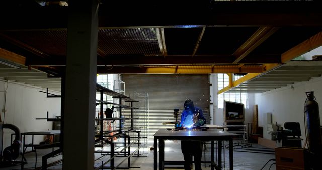 Factory worker wearing a blue welding mask and protective gear, welding at a table in an industrial warehouse. Sparkles are visible, indicating active metalwork. Background includes various pieces of machinery and tools in a spacious, well-lit workshop. Suitable for usage in contexts related to industrial work, metal fabrication, manufacturing processes, and safety gear.