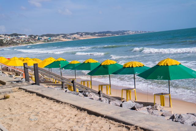 Green and yellow parasols lined up on a sandy beach beside blue ocean waves under a clear sky. Useful for promoting beach destinations, summer vacations, travel blogs and leisure activity advertisements.