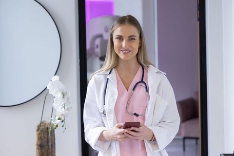 Smiling female doctor standing in modern clinic doorway wearing white coat and pink scrubs