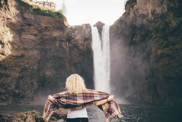 Blonde woman standing in front of a large waterfall, holding a plaid scarf, enjoying the natural beauty and serene environment. Ideal for promoting travel destinations, outdoor activities, relaxation techniques, or adventure tours.