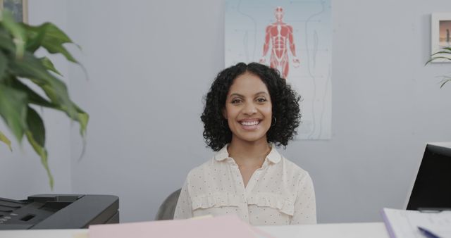 Female receptionist smiling at front desk in medical clinic. Useful for illustrating healthcare services, professional office settings, or healthcare-related websites and materials. Can be used for marketing materials, brochures, or service promotions featuring welcoming staff.