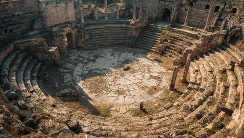 Sunlit ancient amphitheater featuring cracked orchestra, concentric seating and weathered columns
