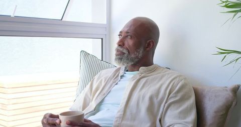 Senior african man relaxing on sofa holding mug and gazing out window