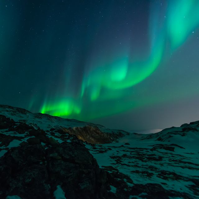 Northern lights create a stunning display over a snow-covered mountain landscape at night. This natural phenomenon, also known as aurora borealis, showcases vibrant green lights dancing in the dark sky. Ideal for use in travel, nature, and science publications to evoke the beauty of the natural world.