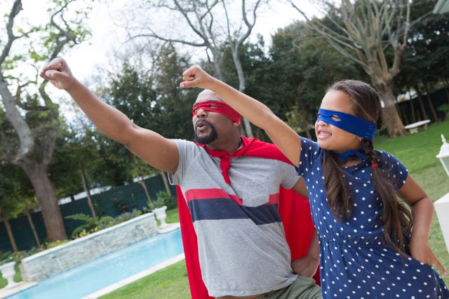 Father and daughter dressed as superheroes, enjoying imaginative play in their backyard. Both are wearing masks and capes, striking heroic poses. Ideal for use in family-oriented content, parenting blogs, advertisements promoting family activities, and articles about childhood imagination and bonding.