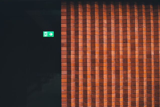Outdoor or indoor views of brick walls with geometric patterns suit content related to architecture, design, or interior decor. This photo shows a minimalist design juxtaposed with an illuminated emergency exit sign, emphasizing the blend of aesthetic and functional elements important for commercial spaces. It can be used for building safety, architectural firms, and interior design projects.