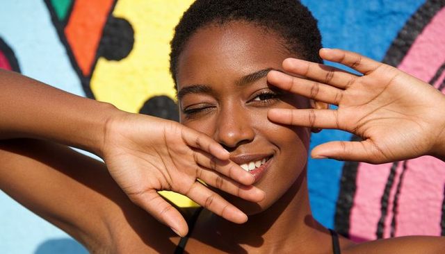 Sunlit smiling woman winking, framing face with hands against colorful urban mural portrait