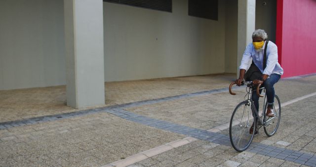 African american senior man wearing face mask riding bicycle in corporate park. hygiene and social distancing during coronavirus covid-19 pandemic.