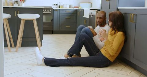 Casual Couple Sitting Comfortably on Kitchen Floor Sipping Hot Drinks