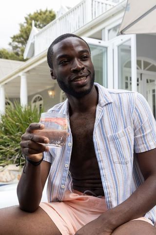 African american man relaxing poolside holding citrus tumbler, summer leisure lifestyle