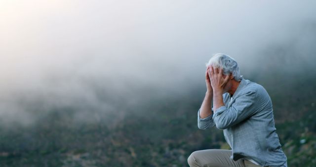 This image depicts an elderly person holding their head in contemplation with a dense fog in the background, creating a serene and thoughtful atmosphere. Ideal for use in articles or adverts addressing themes of reflection, solitude, wellness, aging, mental health, and mindfulness. It is also good for content related to nature, outdoor activities, and tranquil scenery.